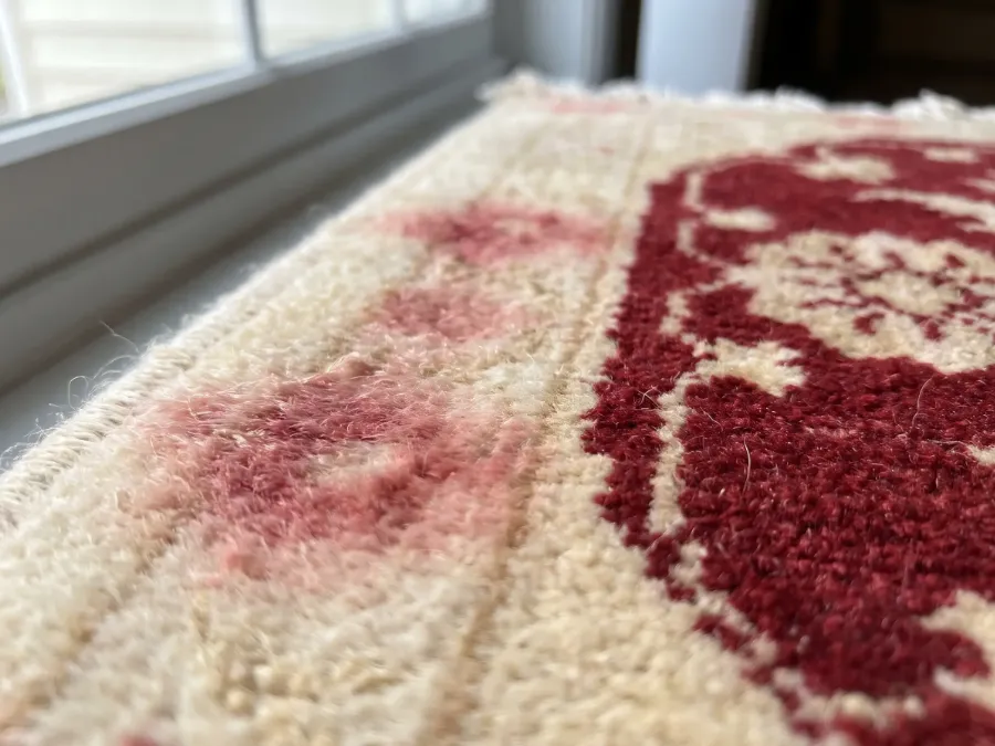 Gros plan d'un tapis d'Orient dont le rouge a coulé dans la bordure ivoire, formant un halo rosé autour des motifs (illustration)